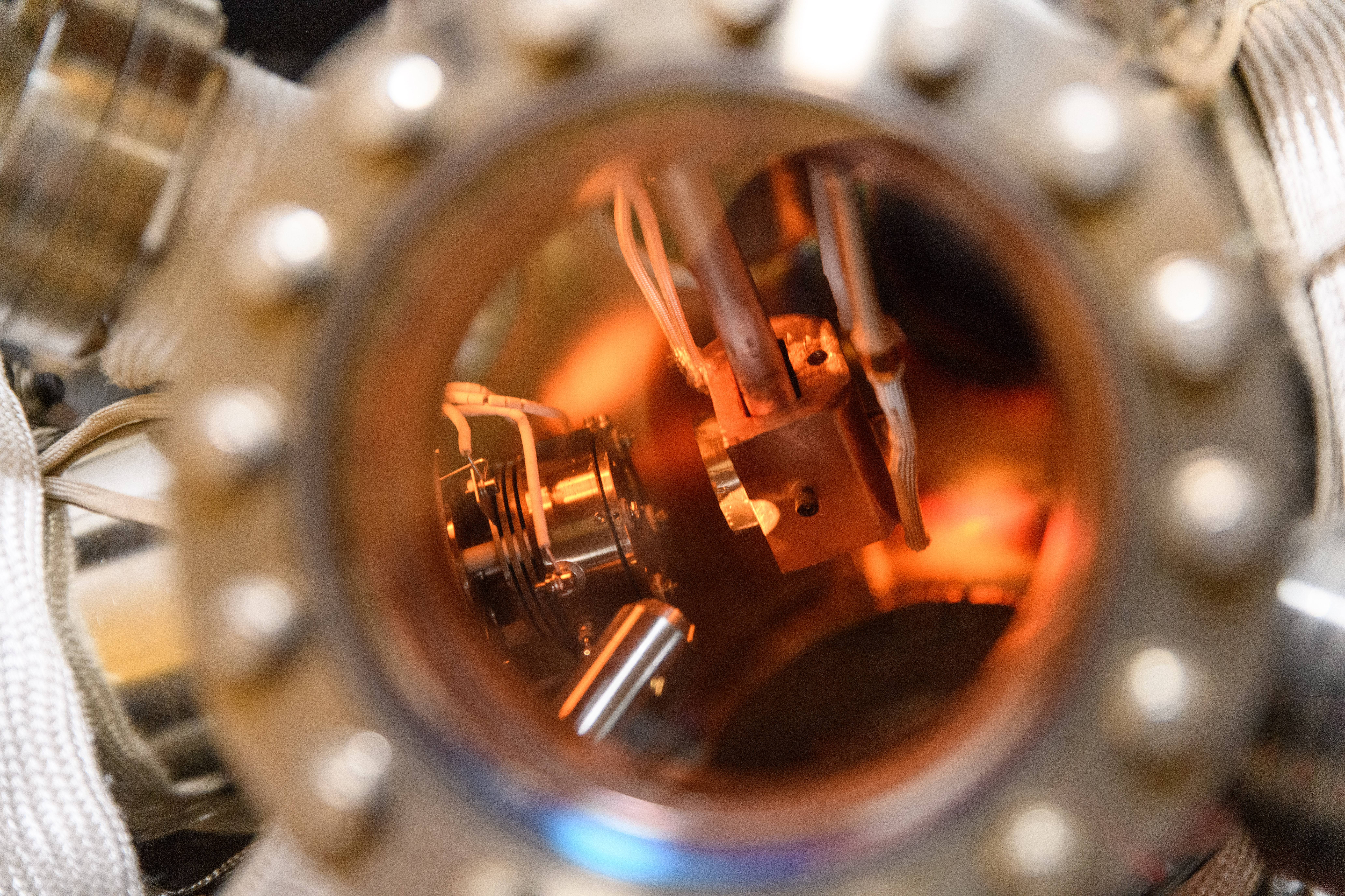 Close-up view through a circular equipment into a laboratory vacuum chamber. Metallic components, cables, and precision instruments are visible inside, illuminated by a warm orange glow. Bolts around the chamber opening frame the image, highlighting the complex experimental equipment used for advanced materials, optics, or physics research.