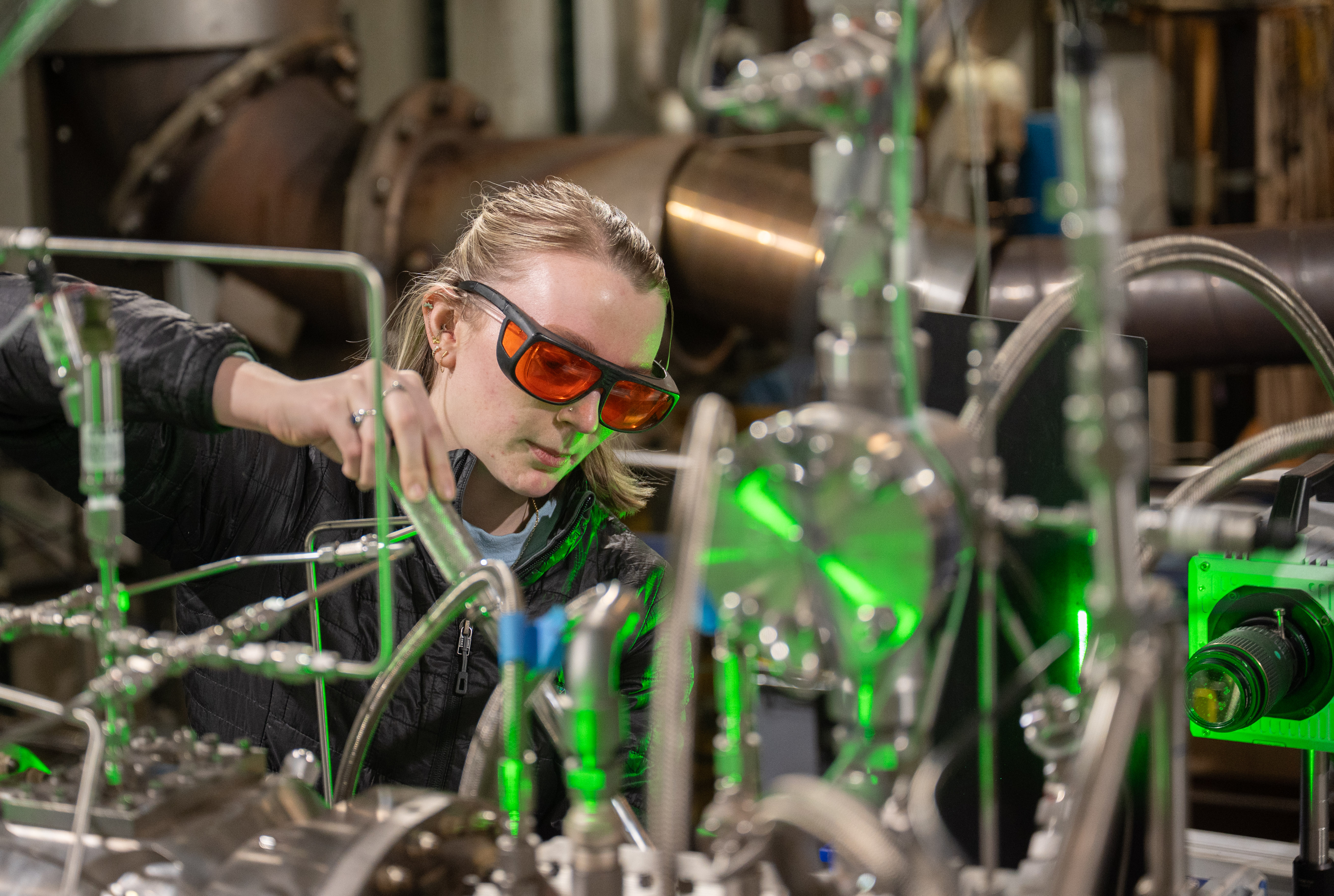 A researcher wearing orange-tinted laser safety glasses adjusts a network of metal pipes, optical components, and sensors in a laboratory. Green laser light shines through the experimental apparatus, with additional scientific equipment visible in the background.