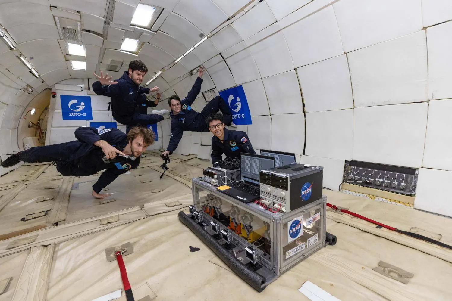 Researchers float in microgravity aboard a Zero-G aircraft beside a NASA-labeled equipment rack containing laptops, electronics, and experimental hardware.