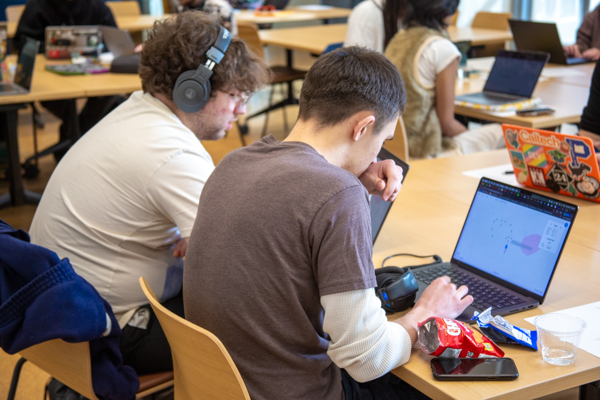 Seated at a long table in the Georgia Tech Library, two participants work on laptops—one screen displaying a node-based audio or visual workflow. Headphones, a notebook, a smartphone, cables, and snack wrappers sit on the tabletop as other attendees work in the background.